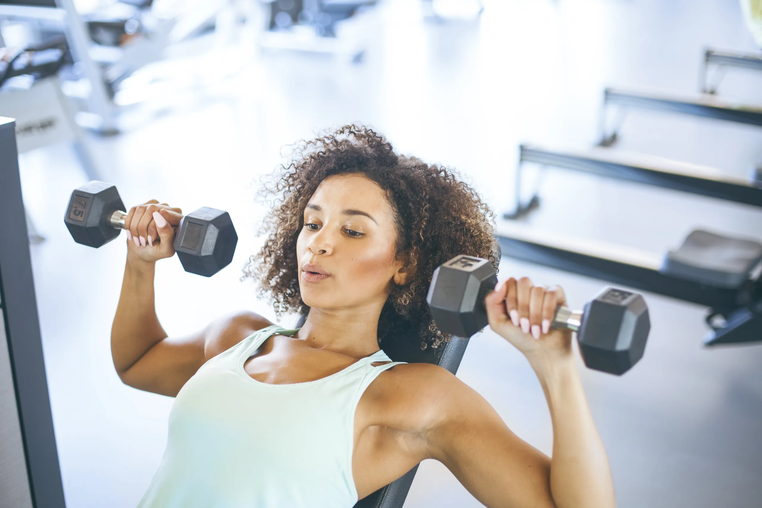 Woman lifting weights as she lays on the bench in a gym. She is wearing a tank top and has curly hair; her lips are pierced as she exhales while exercising.