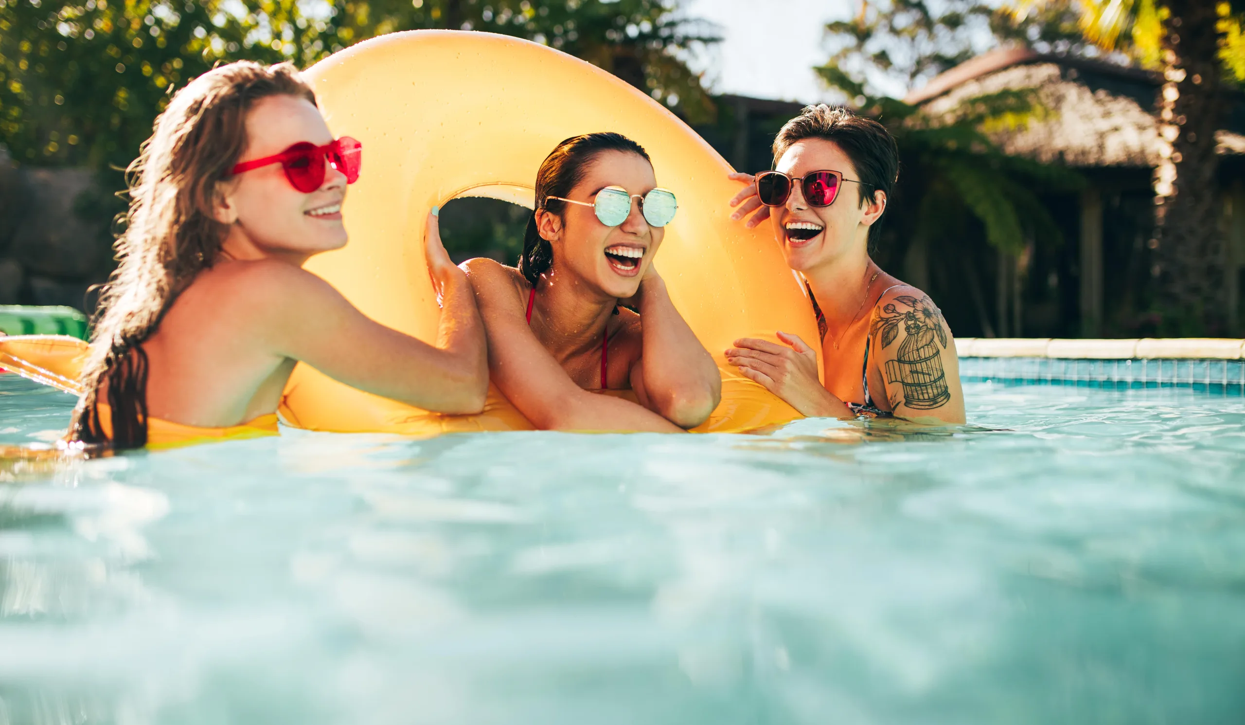 Three young women who are close friends take a selfie with a yellow ring pool float. There is one woman in the middle of the ring. She has gold sunglasses and a red bikini. The woman on the left wearing a mustard yellow swimsuit and has red sunglasses. She is holding the side of the float. The third woman is on the far right and in a blue and white abstract pattern bikini with brown sunglasses, short hair, and a left arm tattoo of a plant with flowers and a bird cage.